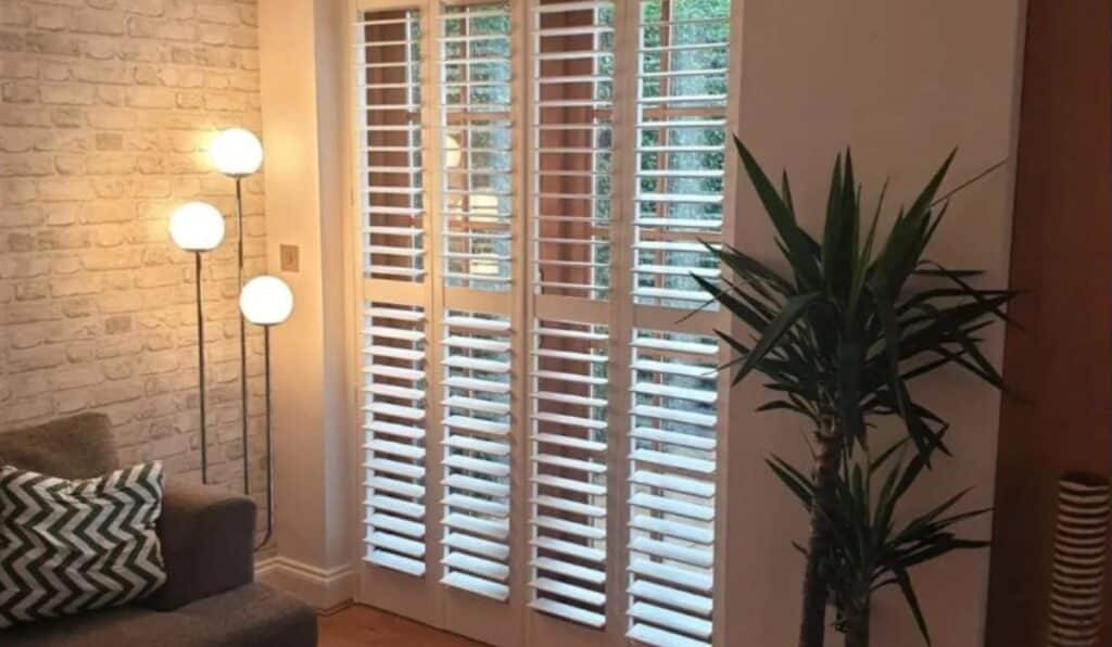 An image showing full height shutters. A 3 light lamp is in the corner for ambient lighting as well as a sofa, coffee table, tall plant and scratching post.
