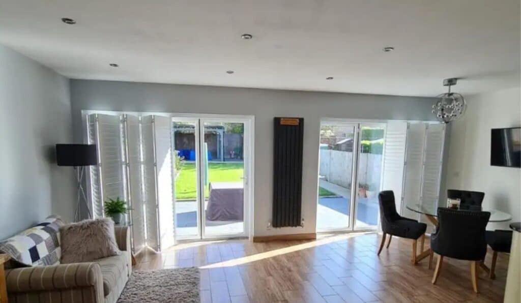 a large room with tracked shutters, a sofa to the left and a dining table to the right.